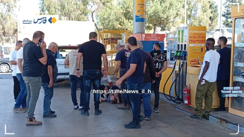 A man lies on the ground at a gas station as several bystanders stand around him, with Hebrew and Arabic signage visible.