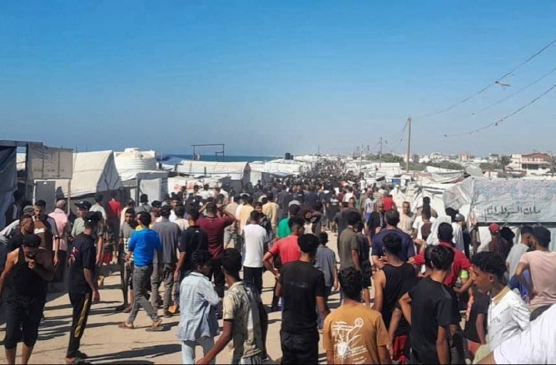 A large crowd of people, many carrying belongings, walks through a dusty area lined with tents near a coastline.