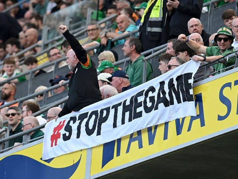Fans in green attire stand in stadium seating, with one man raising his fist behind a banner reading "#STOPTHEGAME" above a yellow advertising hoarding.
