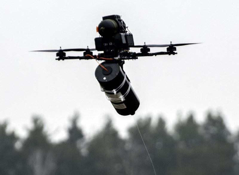 A large multi-rotor drone equipped with a mounted camera and a cylindrical payload hovers in mid-air against a pale sky, with a treeline visible below.