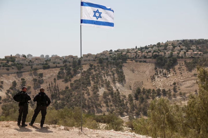Two uniformed personnel stand on a hillside overlooking a dry, terraced valley with residential buildings, while a large Israeli flag waves on a pole in the foreground.