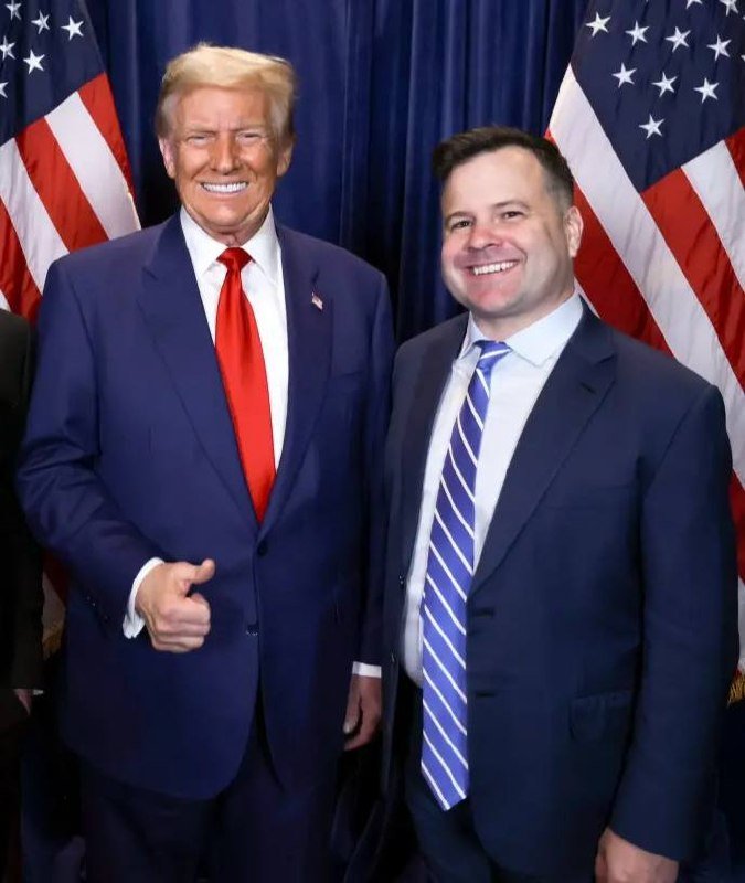 Two smiling men in dark suits stand side-by-side in front of two American flags against a dark blue backdrop.