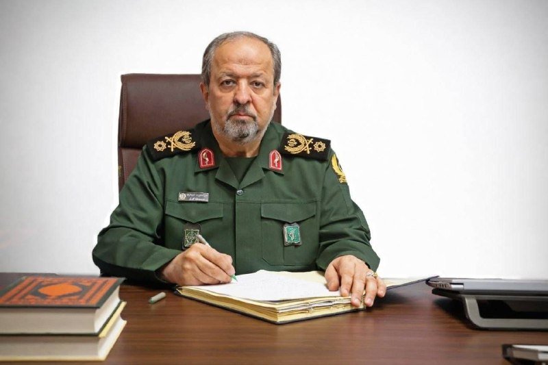 A man with a gray beard, wearing an olive-green military uniform with insignia, sits at a desk signing a document, with stacked books and a laptop nearby.