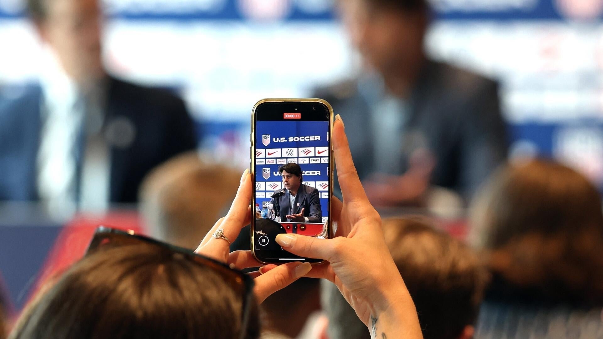 A person holds a smartphone recording a man speaking at a podium during a U.S. Soccer press conference.