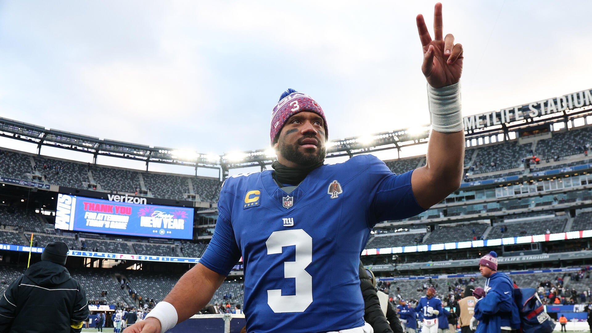 A football player in a blue jersey with the number 3 raises two fingers while walking on a stadium field, with a scoreboard displaying a "Thank You" message behind him.