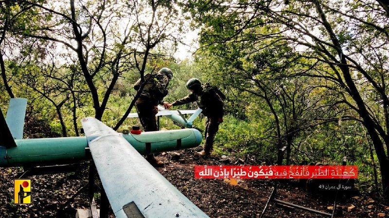 Two helmeted figures in tactical gear stand among trees, examining a downed light-blue unmanned aerial vehicle with visible wings and a fuselage, alongside Arabic text overlay.
