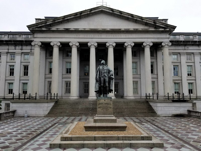 A bronze statue of a man in a long coat stands on a stone pedestal in front of a neoclassical building labeled "THE TREASURY DEPARTMENT," featuring tall Ionic columns and a grand staircase.