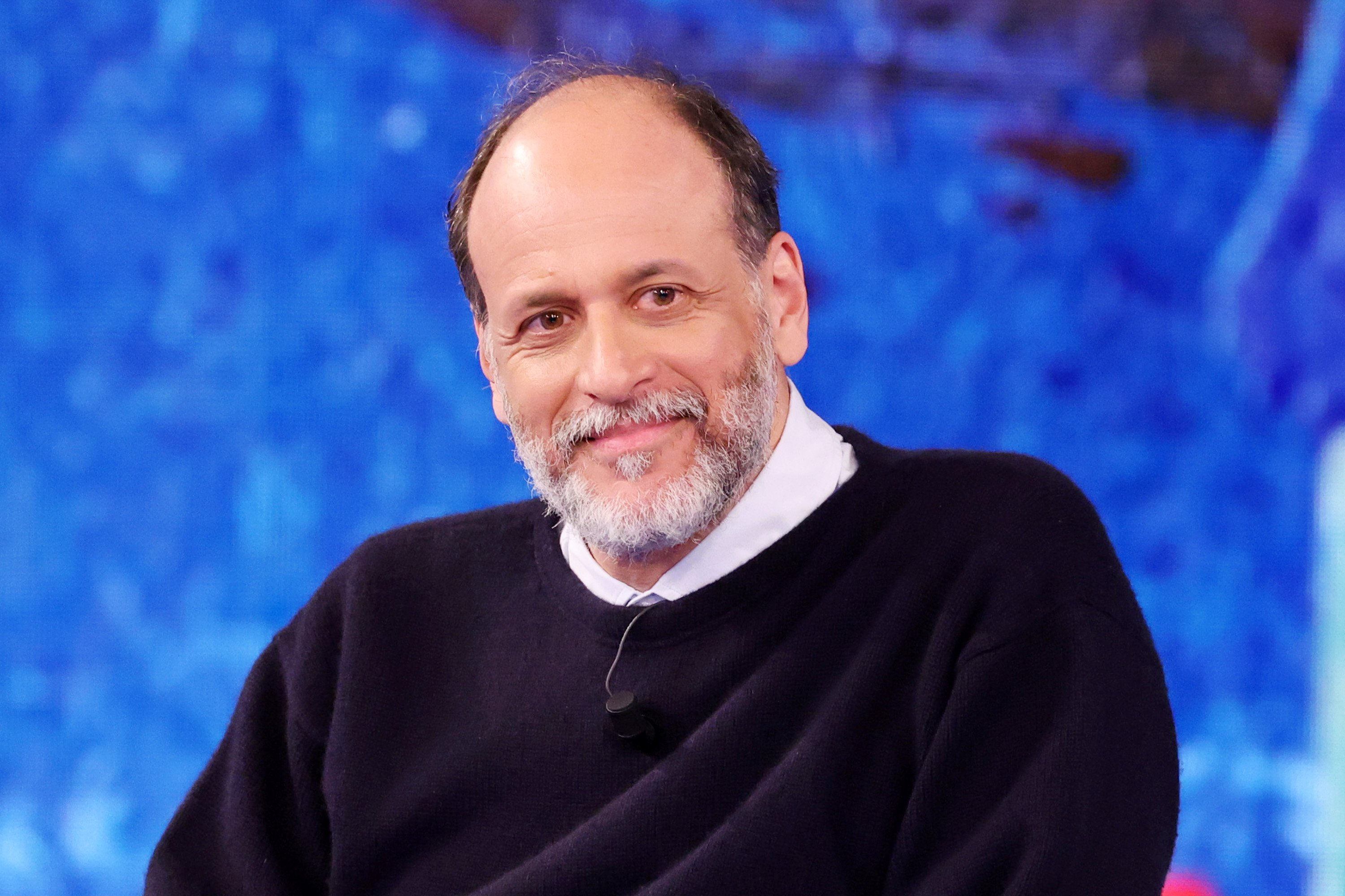 A smiling man with a gray beard, wearing a black sweater over a white collared shirt and a lapel microphone, stands before a blue backdrop.