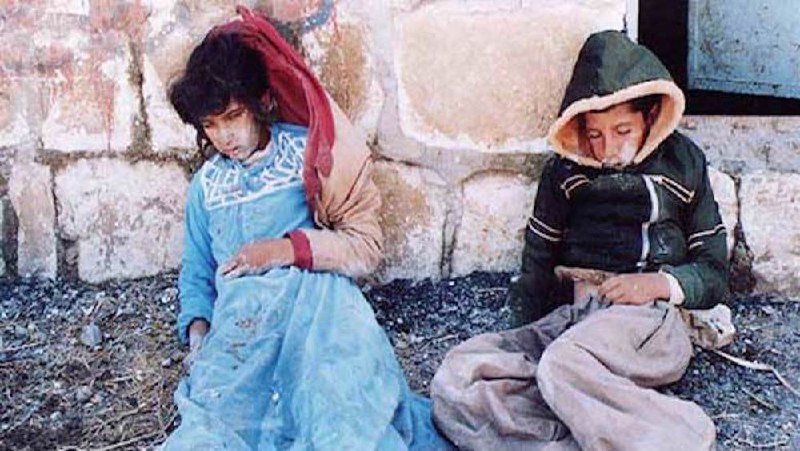 Two children in hooded jackets sit slumped against a weathered stone wall, their heads bowed.