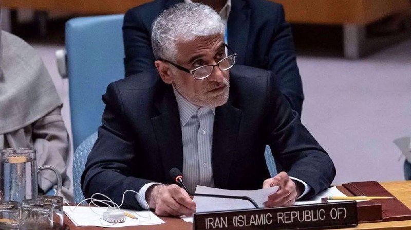 A gray-bearded man wearing glasses and a dark suit writes on documents at a desk labeled "IRAN (ISLAMIC REPUBLIC OF)" in a formal meeting setting.