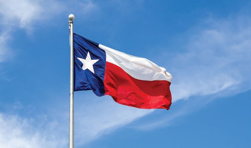 The Texas state flag waves on a flagpole against a blue sky with scattered white clouds.