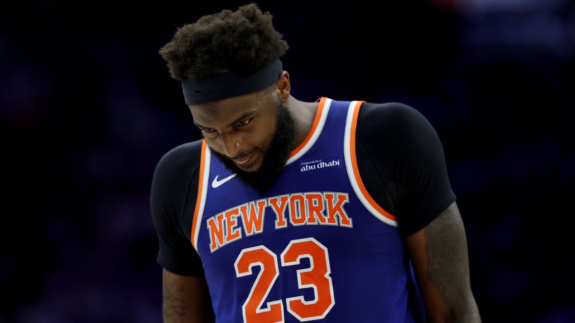 A bearded basketball player wearing a blue "New York" jersey with the number 23 and a black headband looks downward against a dark background.