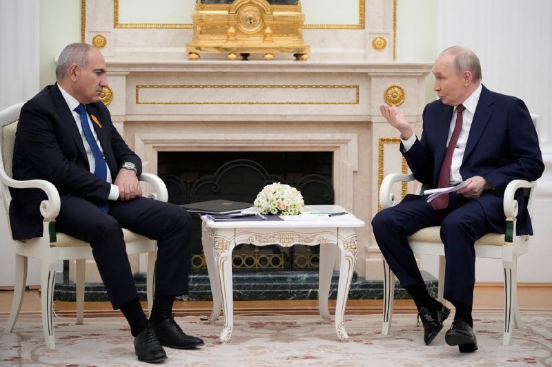 Two men in dark suits sit in ornate chairs facing each other in a formal room, with one gesturing while speaking during a meeting beside a small table with flowers.