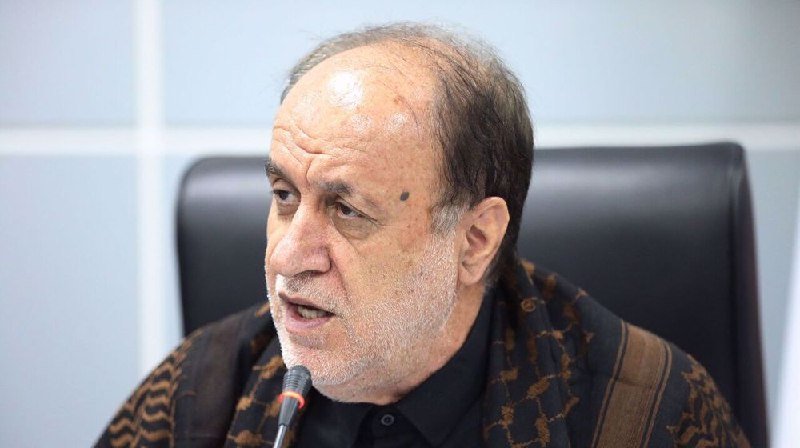 An older man with a graying beard, wearing a dark patterned shawl over a black shirt, speaks into a microphone while seated in a black office chair.