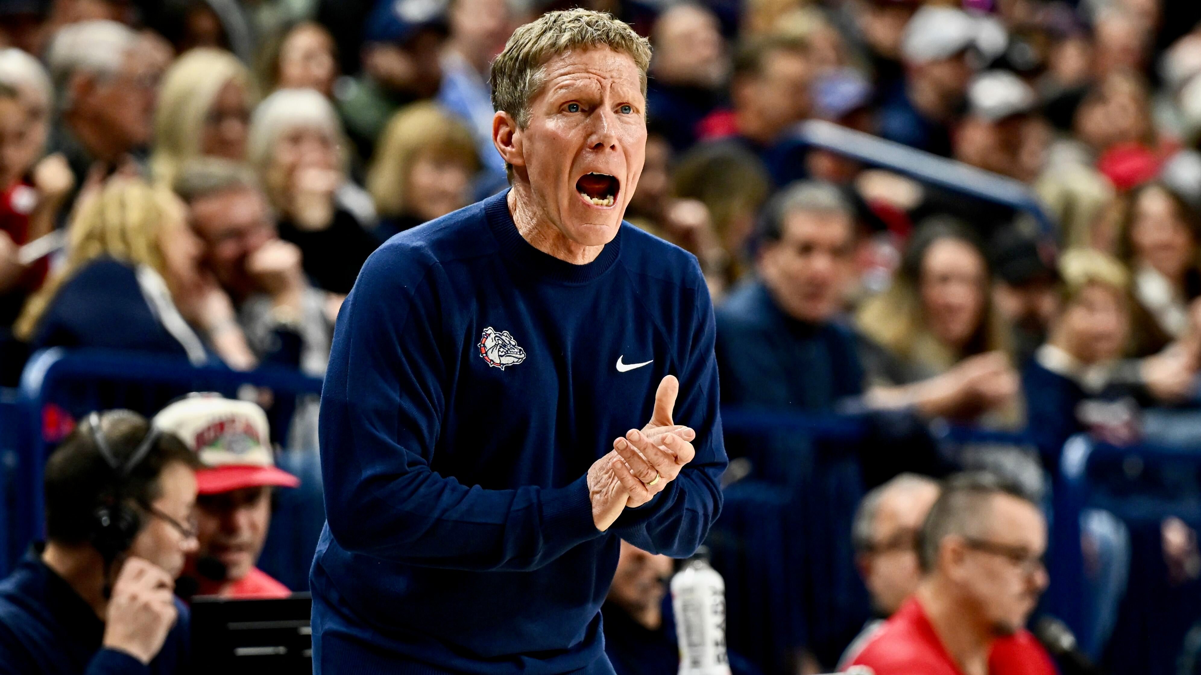 A blonde coach in a navy Bulldogs sweatshirt shouts and claps his hands from the sideline during a basketball game, with a crowd visible behind him.