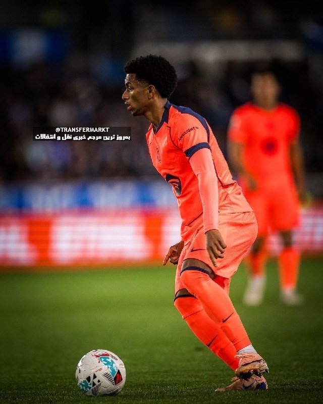 A soccer player in an orange uniform stands on a grass pitch, poised beside a ball, with a blurred teammate and stadium crowd visible in the background.