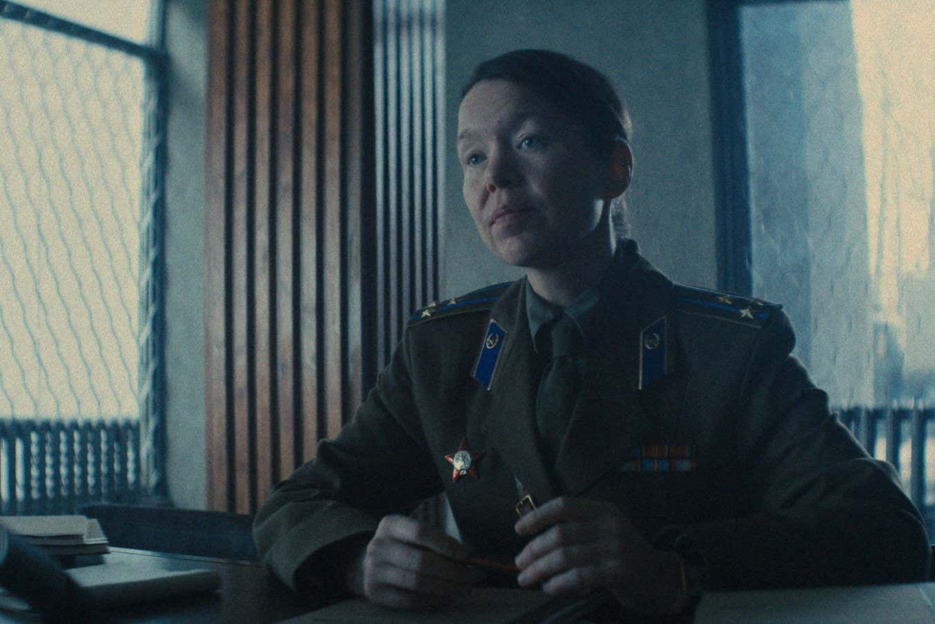 A woman in a dark military uniform with shoulder boards, collar insignia, and a star medal sits at a desk holding a pen, with a window and radiator visible behind her.