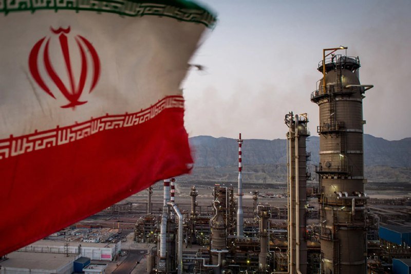 The Iranian flag waves in the foreground of an industrial refinery complex with tall towers and pipes set against a mountainous backdrop.