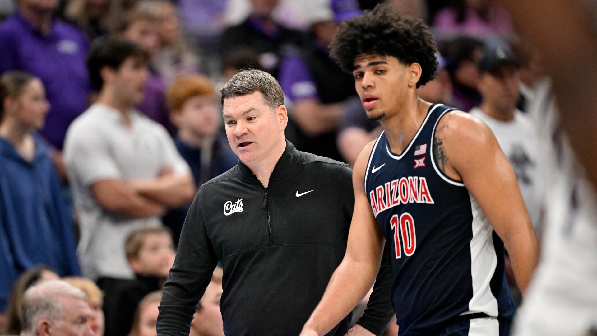 A man in a black "Cats" jacket walks alongside an Arizona basketball player wearing jersey number 10, with a crowd visible in the background.