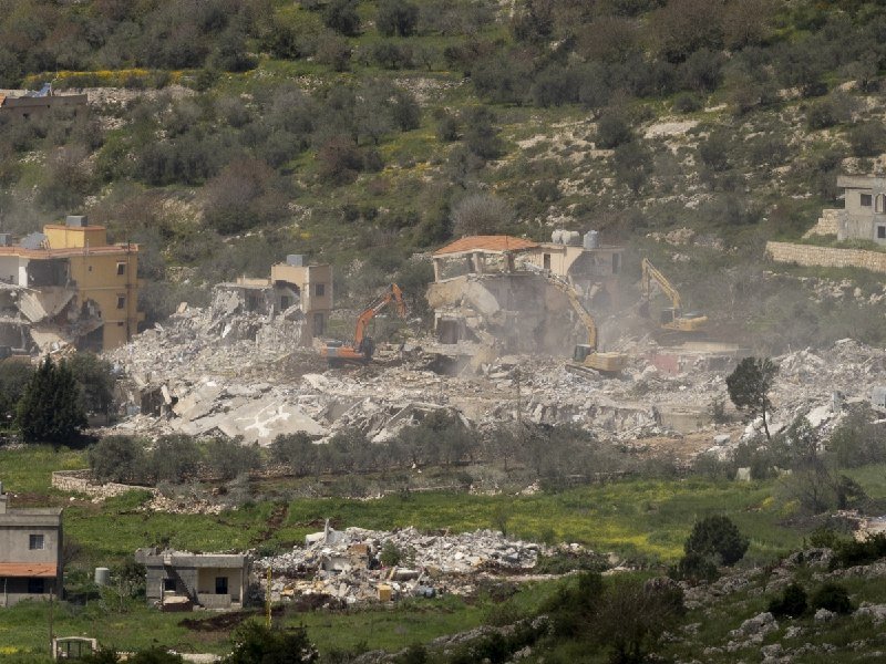Excavators and workers demolish rubble-strewn buildings amid green, terraced hillsides.