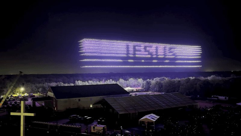 Hundreds of illuminated drones form a glowing rectangular display above a rural landscape featuring barns, solar panels, and an illuminated cross at night.