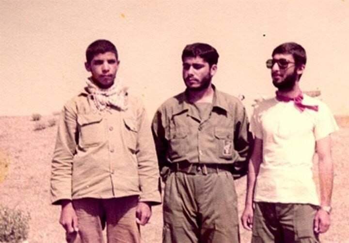 Three young men stand side by side outdoors in a dry, open landscape, wearing military-style and casual clothing in a faded color photograph.