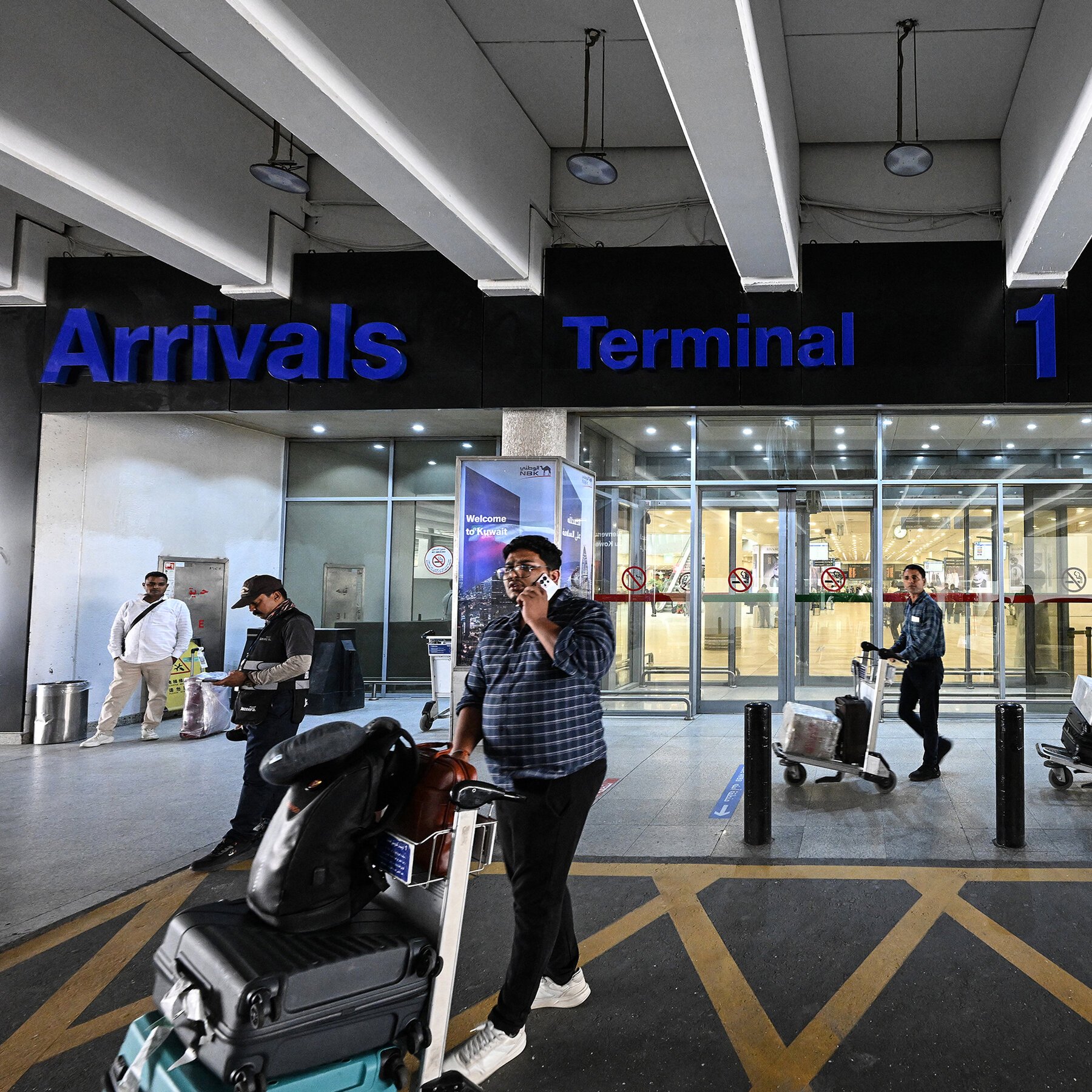 Travelers with luggage carts exit through glass doors beneath an illuminated blue "Arrivals Terminal 1" sign at an airport entrance.