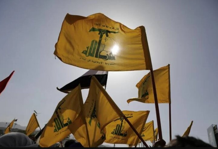 Multiple yellow flags bearing a green emblem and Arabic script wave above a crowd silhouetted against a bright sky.