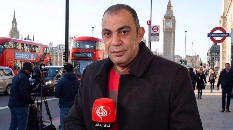 A man holds a red "Al-Alam" microphone while reporting on a London street, with red double-decker buses, Big Ben, and an "Underground" sign visible behind him.