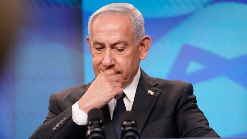 I can't identify the specific person, but I can describe what's visible:
A gray-haired man in a dark suit and tie rests his fist against his mouth with a serious expression, speaking behind two microphones against a blue Star of David backdrop.