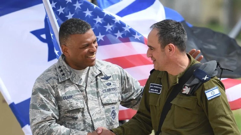 Two smiling military officers—one in a U.S. Air Force uniform, the other in an Israeli Air Force uniform with Hebrew text—shake hands in front of U.S. and Israeli flags.