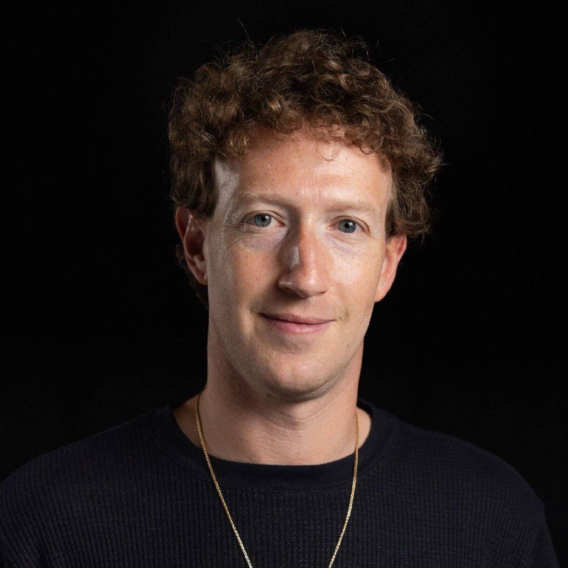 A man with curly reddish-brown hair, wearing a dark sweater and thin gold chain, smiles at the camera against a black background.