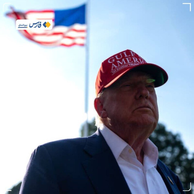 An older man wearing a red "GULF OF AMERICA" cap and dark blazer stands outdoors with a U.S. flag waving in the background.