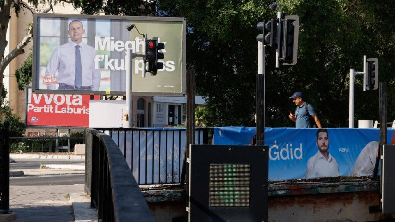 A pedestrian in a blue cap walks past election billboards featuring men in suits and party text including "PN," "Partit Laburista," and "Gdid."
