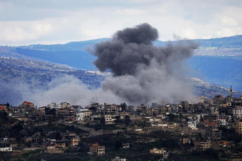 Dark smoke rises above a densely built hillside town surrounded by mountains under a cloudy sky.