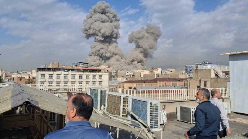 Two large gray smoke plumes rise above a city skyline as several men stand on a rooftop watching.