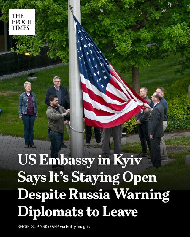 A group of people lower an American flag from a flagpole in a grassy, tree-lined outdoor setting, with overlaid text reading "US Embassy in Kyiv Says It's Staying Open Despite Russia Warning Diplomats to Leave."