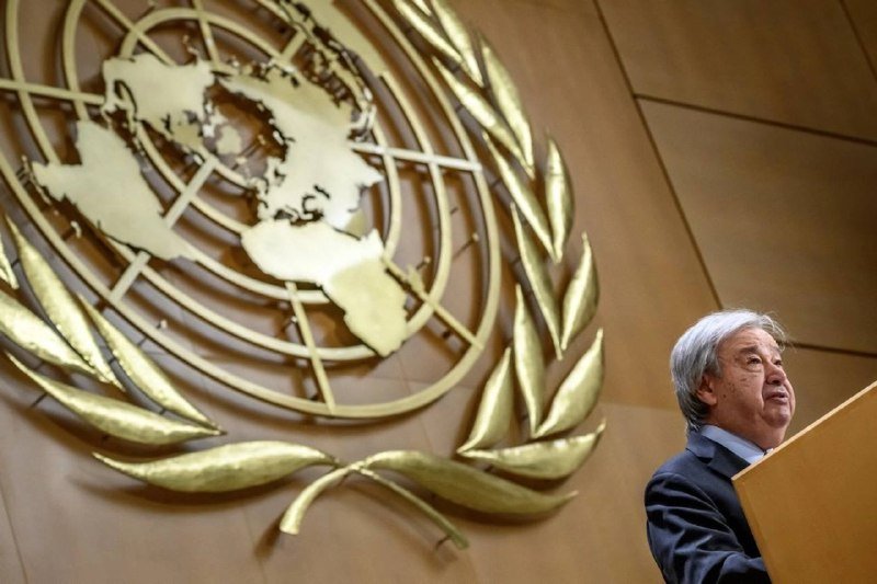 A gray-haired man in a dark suit speaks at a podium beside a large gold United Nations emblem mounted on a wall.