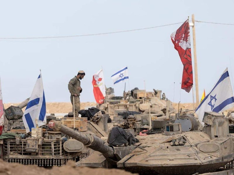 A soldier stands atop a tank in a desert, surrounded by other armored vehicles and multiple Israeli flags.