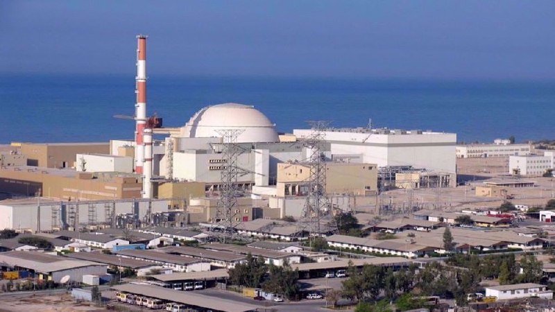 A nuclear power plant with a domed reactor building and red-and-white striped smokestack stands along a coastline, surrounded by industrial structures, power lines, and warehouse facilities.