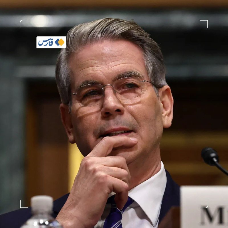 A gray-haired man in a suit and glasses sits thoughtfully with his hand near his chin before a microphone, with a partially visible nameplate.