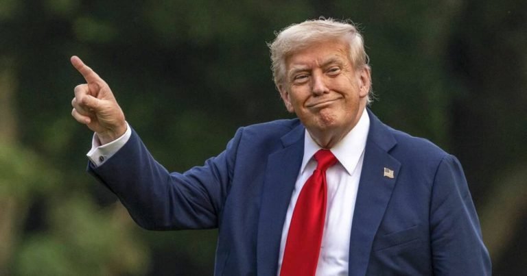A blond man in a navy suit, white shirt, and red tie stands outdoors against a blurred green background, pointing upward with his right hand and smiling.