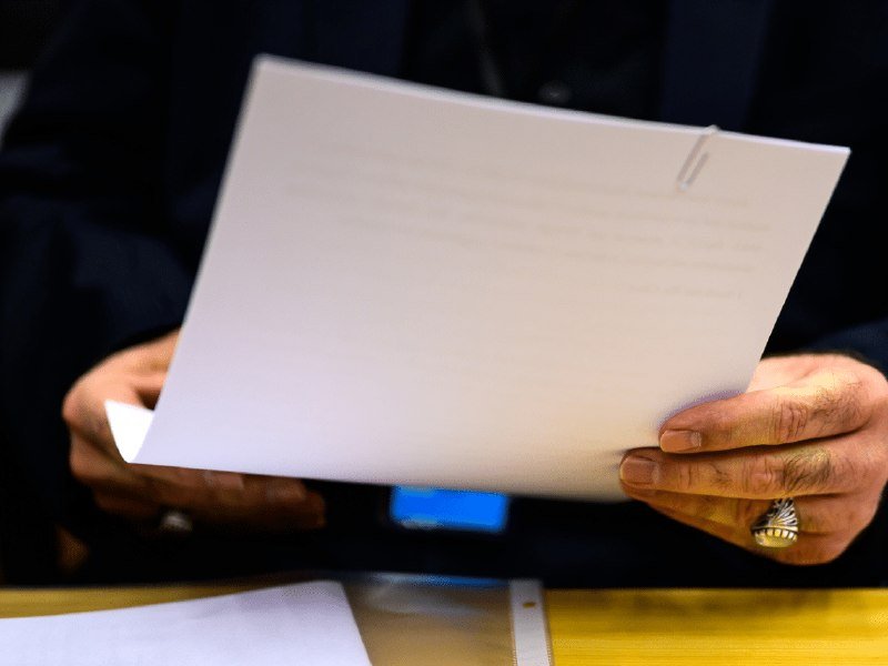 A person wearing a dark shirt holds a white document with both hands, revealing a gold ring on their finger.