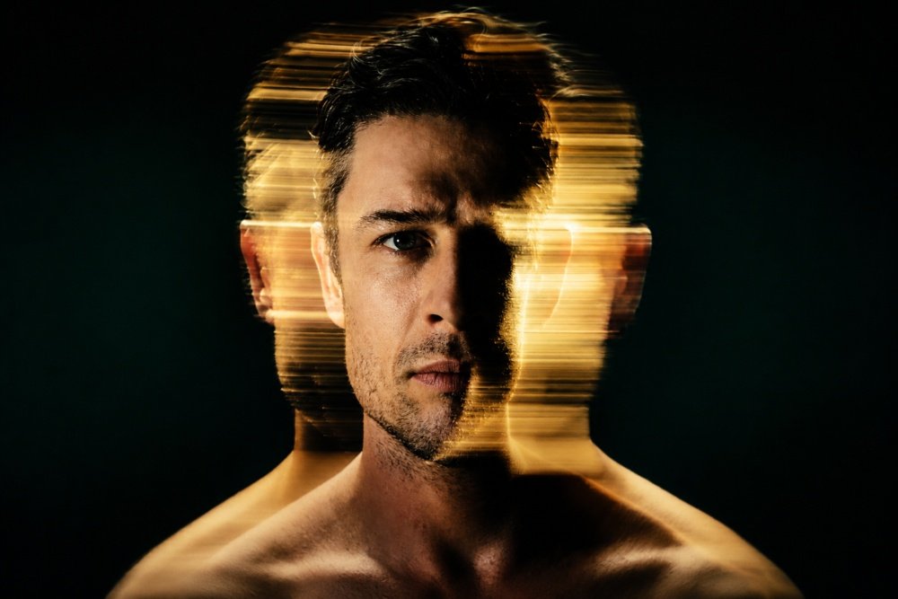 A shirtless man stares forward against a black background, with streaks of golden light and motion-blurred duplicates of his head layered behind his face.