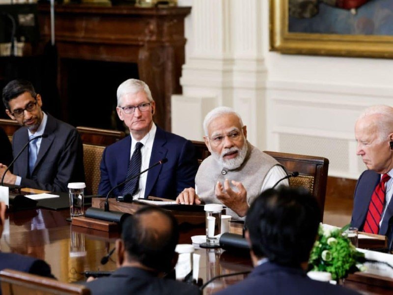 Four men in business attire sit at a long conference table with microphones, water glasses, and papers, engaged in discussion in a formal room with wood paneling and a fireplace.