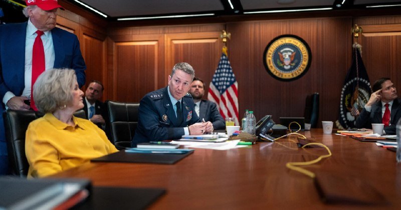 A man in military uniform sits at a wood conference table beside an American flag, with the Presidential Seal on the wall behind him, while several others are seated or standing nearby.