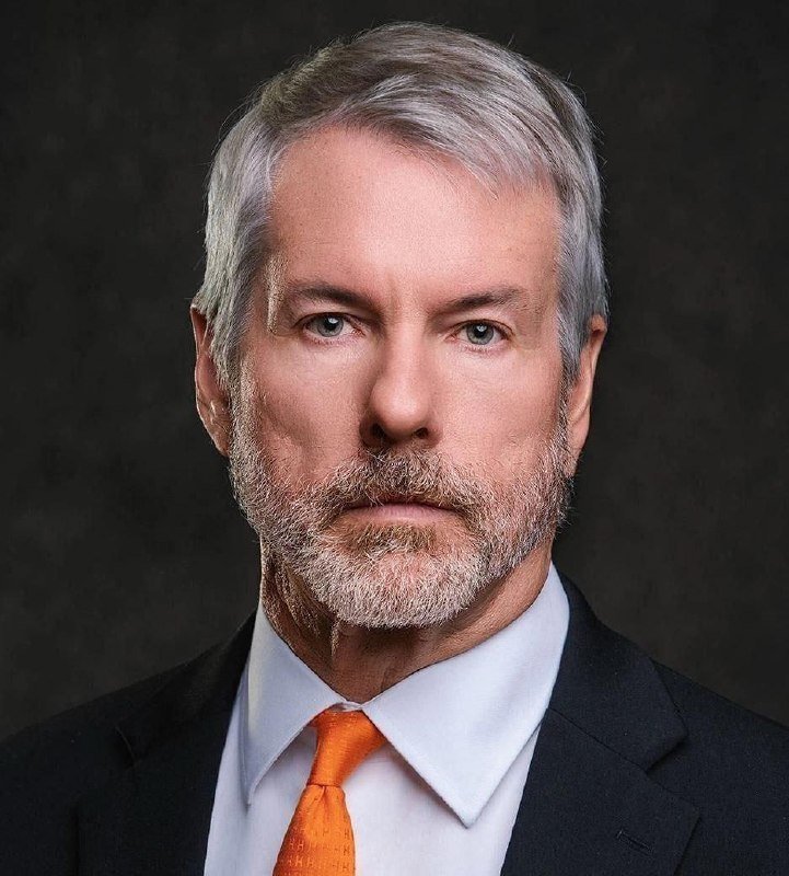 A gray-haired, bearded man in a dark suit, white shirt, and orange tie poses for a formal portrait against a dark background.