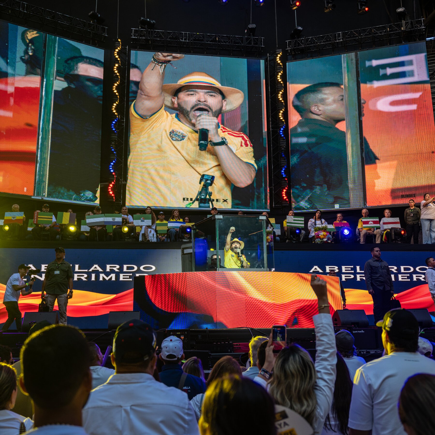 A man in a yellow jersey and hat speaks into a microphone at a podium during a large indoor rally, with his image displayed on giant screens above a seated crowd.
