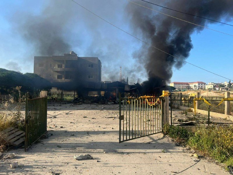 Thick black smoke rises from a burning structure behind an open metal gate, with charred debris scattered across a sandy lot and buildings visible in the background.
