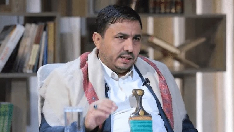 A man wearing a white shirt and patterned scarf speaks while seated in front of bookshelves, with a traditional dagger and a glass of water visible nearby.